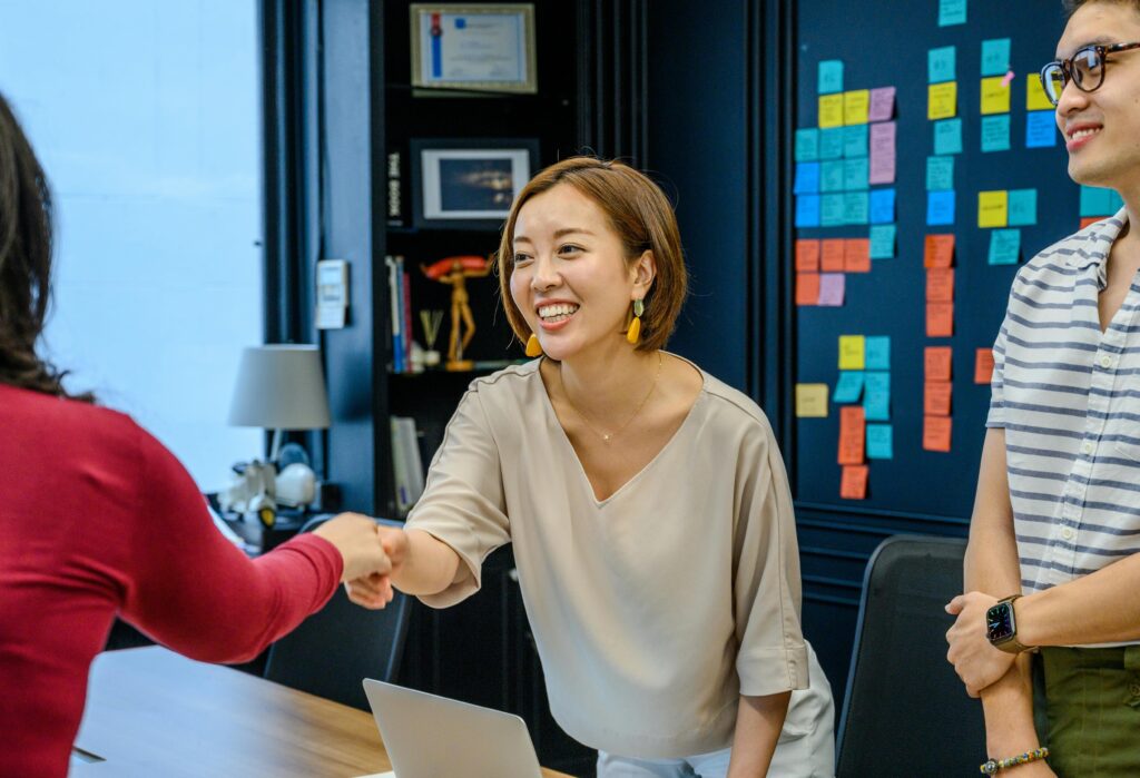 Three colleagues in a meeting room, with a woman smiling and shaking hands.